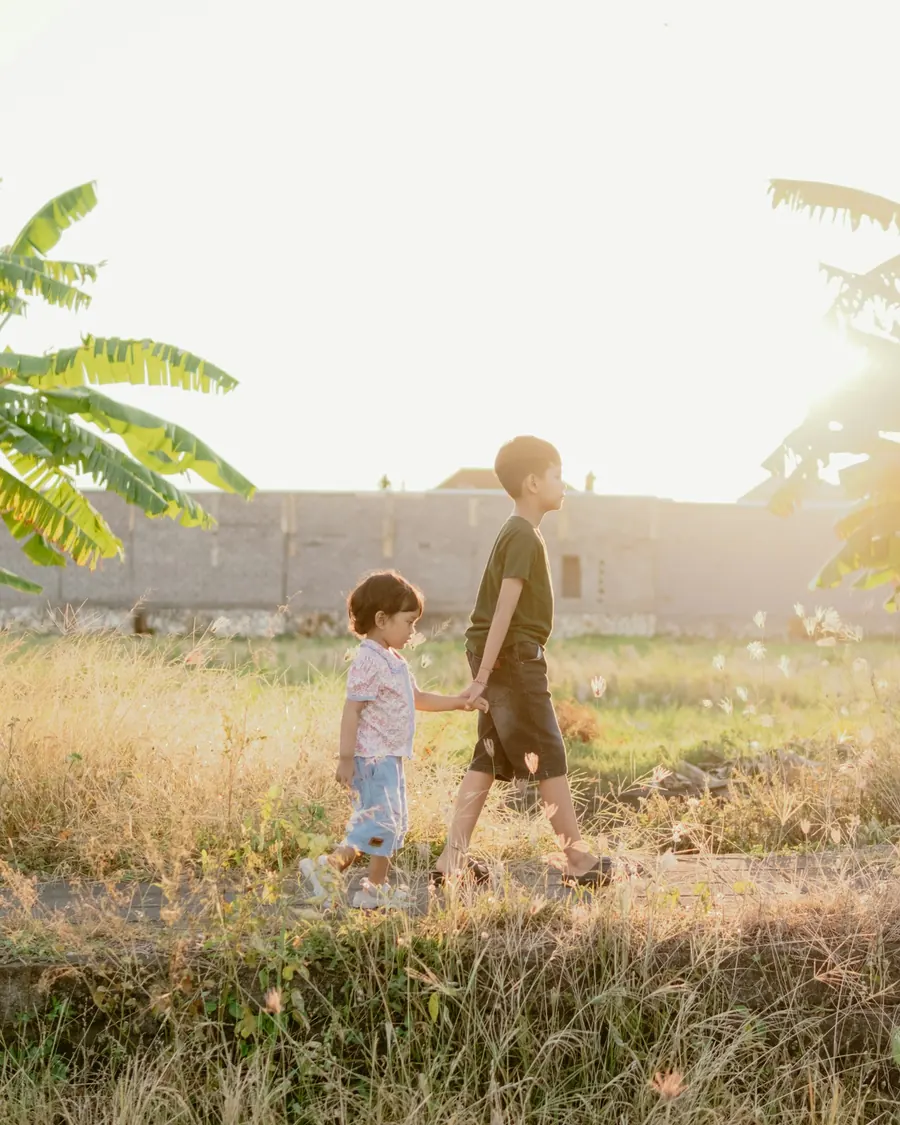 Two young children holding hands as they walk through tall golden grass in evening light